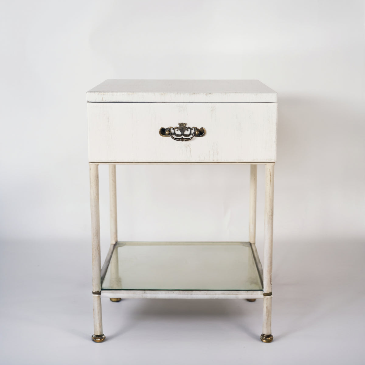 Front view of the white Damme bedside table with drawer, ornate handle and glass shelf, on a white background