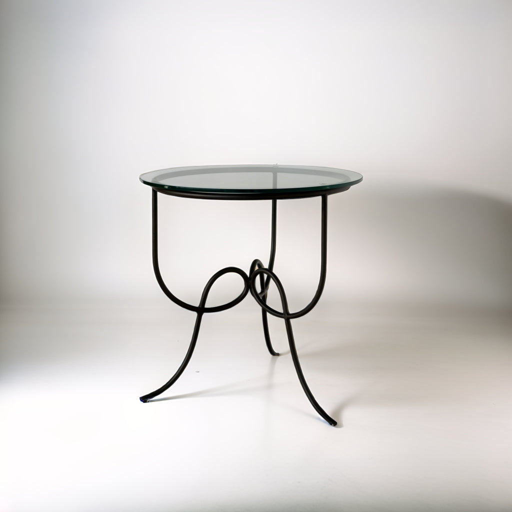 Entony bedside table with round glass top and gracefully scrolled black metal base against a light background